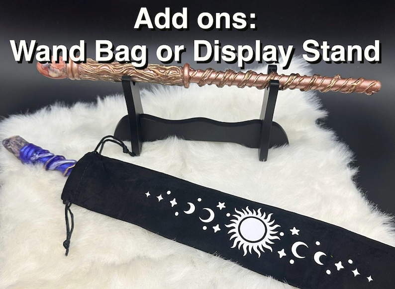 K&ouml;nnte beinhalten: Ein kupferfarbener Zauberstab mit Spiralmuster, ausgestellt auf einem schwarzen St&auml;nder. Eine schwarze Zauberstabtasche mit einem Himmelsdesign mit Monden, Sternen und einer Sonne ist ebenfalls vorhanden. Der Text "Add ons: Wand Bag or Display Stand" steht oben.