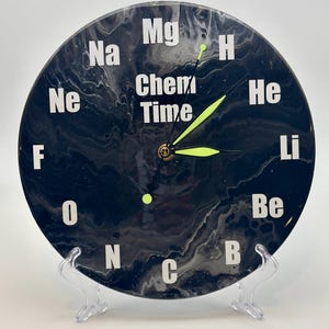 May include: A black and white clock with a marbled design. The clock face features the words "Chem Time" and the chemical symbols for various elements, including Na, Mg, H, He, Li, Be, F, O, N, C, and B.