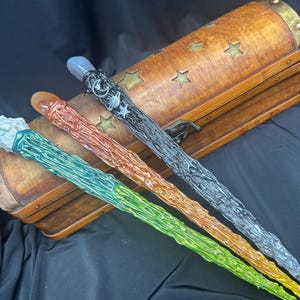K&ouml;nnte beinhalten: Vier farbenfrohe Zauberst&auml;be mit strukturierten Designs und Edelsteinspitzen werden gezeigt. Die Zauberst&auml;be sind gr&uuml;n, orange, silber und schwarz. Im Hintergrund befindet sich eine Holzkiste mit Sternaussparungen und Messingakzenten.