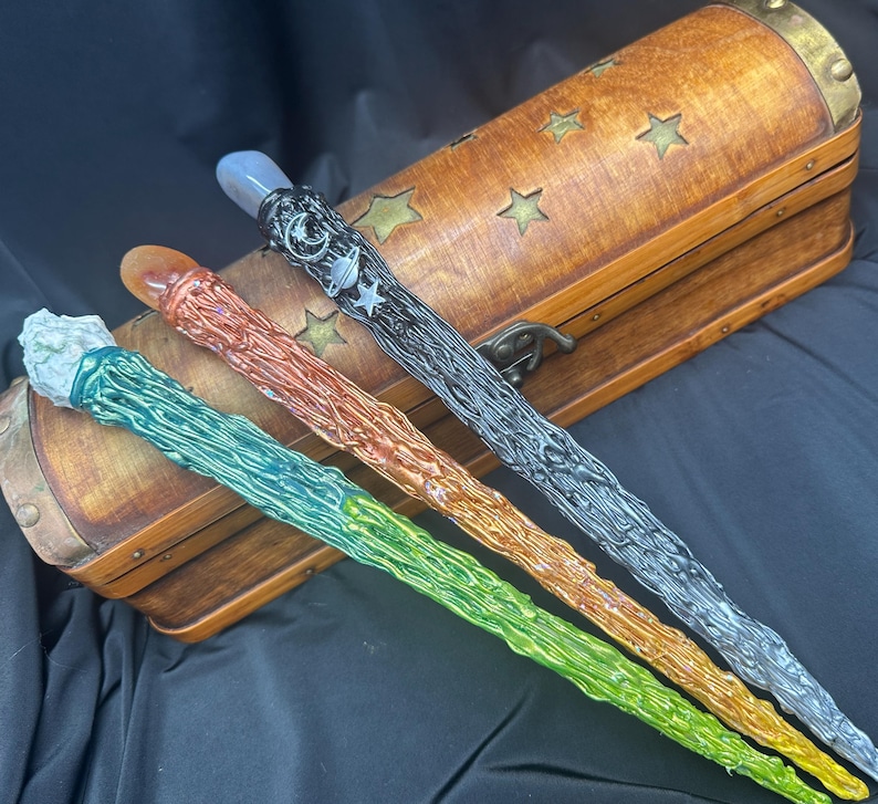 K&ouml;nnte beinhalten: Vier farbenfrohe Zauberst&auml;be mit strukturierten Designs und Edelsteinspitzen werden gezeigt. Die Zauberst&auml;be sind gr&uuml;n, orange, silber und schwarz. Im Hintergrund befindet sich eine Holzkiste mit Sternaussparungen und Messingakzenten.