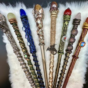 K&ouml;nnte beinhalten: Eine Sammlung von acht handgefertigten Zauberst&auml;ben mit einzigartigen Designs. Jeder Zauberstab hat ein anderes Farbschema, einen Edelstein und dekorative Elemente, darunter Drachen, Monde und aufwendige Metallarbeiten. Die Zauberst&auml;be werden auf einer wei&szlig;en, flauschigen Oberfl&auml;che pr&auml;sentiert.