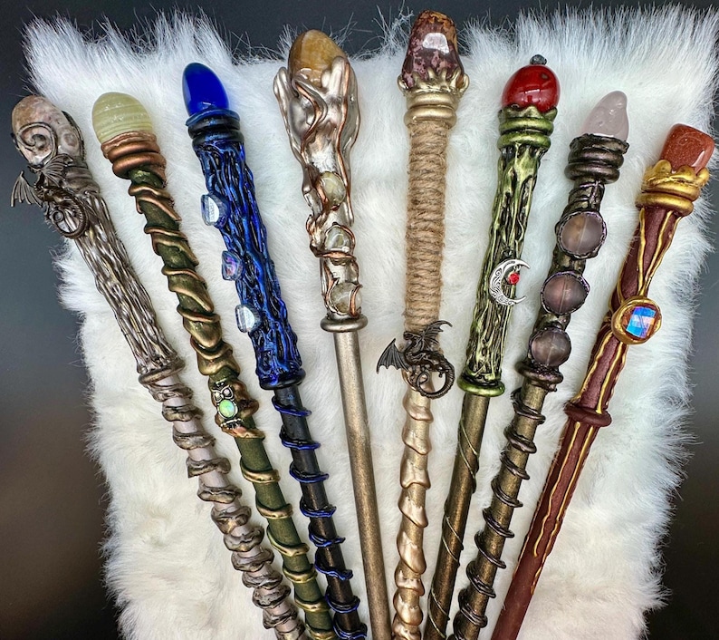 K&ouml;nnte beinhalten: Eine Sammlung von acht handgefertigten Zauberst&auml;ben mit einzigartigen Designs. Jeder Zauberstab hat ein anderes Farbschema, einen Edelstein und dekorative Elemente, darunter Drachen, Monde und aufwendige Metallarbeiten. Die Zauberst&auml;be werden auf einer wei&szlig;en, flauschigen Oberfl&auml;che pr&auml;sentiert.