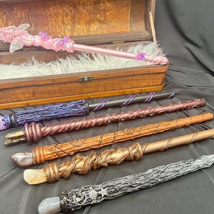 K&ouml;nnte beinhalten: Eine Sammlung handgefertigter Zauberst&auml;be in verschiedenen Farben und Designs. Die Zauberst&auml;be bestehen aus Holz und Harz mit Details wie Kristallen, Wirbeln und floralen Akzenten. Ein Zauberstab ist rosa mit einem Schmetterlingsdesign, ein anderer ist silberfarben mit Sternendetails.