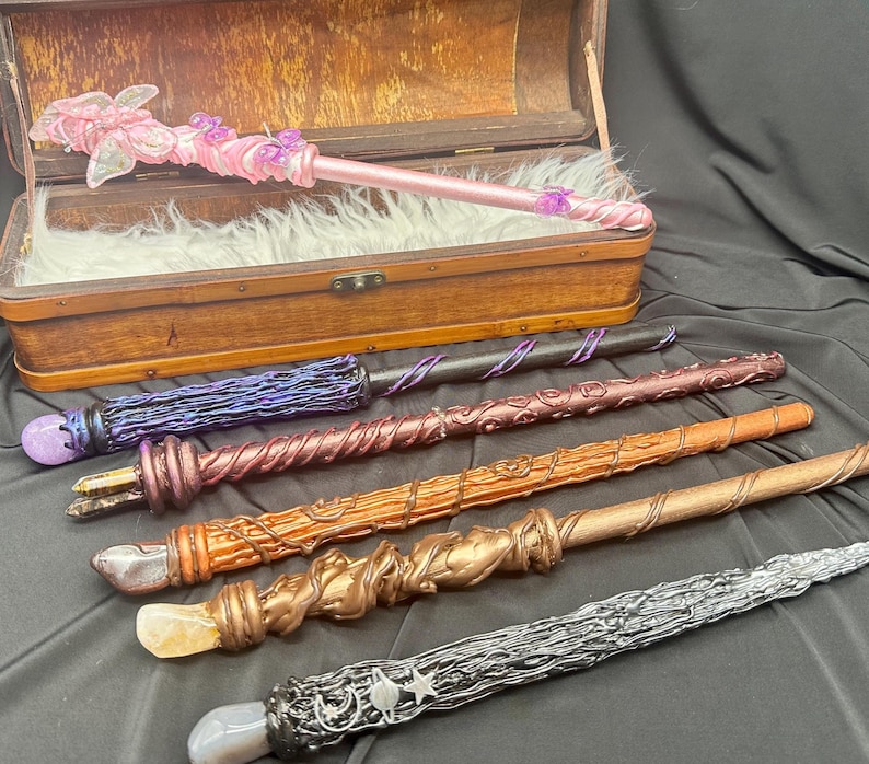 K&ouml;nnte beinhalten: Eine Sammlung handgefertigter Zauberst&auml;be in verschiedenen Farben und Designs. Die Zauberst&auml;be bestehen aus Holz und Harz mit Details wie Kristallen, Wirbeln und floralen Akzenten. Ein Zauberstab ist rosa mit einem Schmetterlingsdesign, ein anderer ist silberfarben mit Sternendetails.