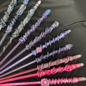 K&ouml;nnte beinhalten: Eine Sammlung handgefertigter Zauberst&auml;be in verschiedenen Farben, darunter Blau, Lila und Rosa. Jeder Zauberstab hat ein einzigartiges Design mit Kristallakzenten und dekorativen Verzierungen. Die Zauberst&auml;be sind auf einem schwarzen Hintergrund angeordnet.