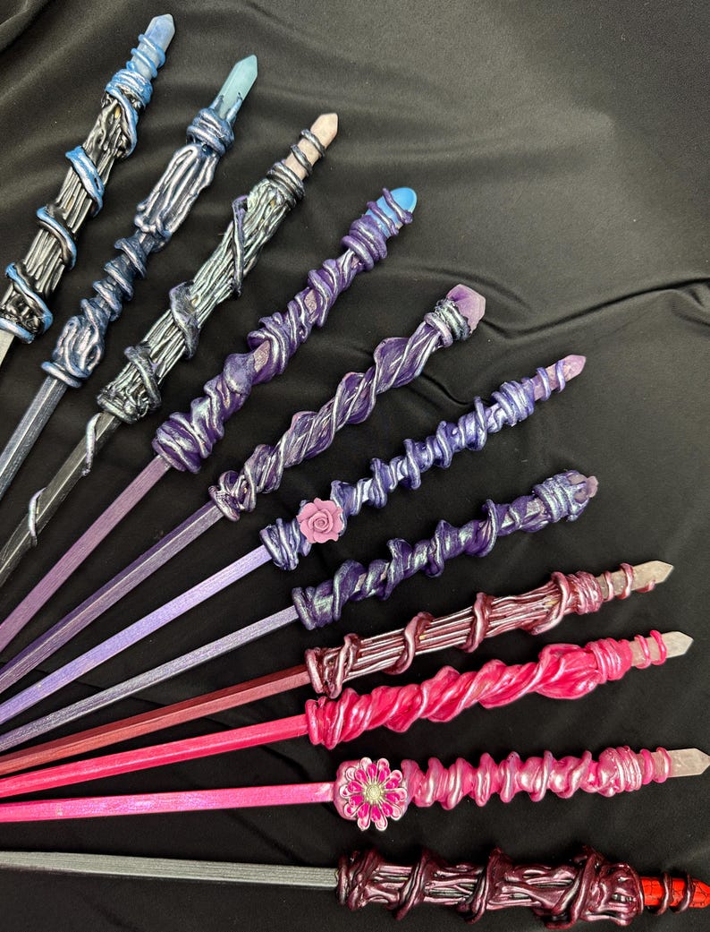 K&ouml;nnte beinhalten: Eine Sammlung handgefertigter Zauberst&auml;be in verschiedenen Farben, darunter Blau, Lila und Rosa. Jeder Zauberstab hat ein einzigartiges Design mit Kristallakzenten und dekorativen Verzierungen. Die Zauberst&auml;be sind auf einem schwarzen Hintergrund angeordnet.