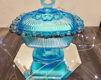 May include: A vintage, light blue glass candy dish with a lid. The dish has a ribbed design and is adorned with decorative elements. It sits on a mirrored, hexagonal base. The lid features a loop handle.