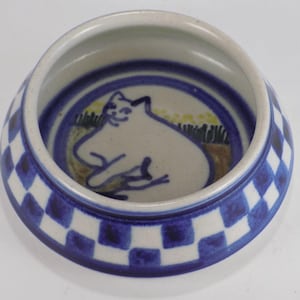 Könnte beinhalten: Keramik-KatzenNapf mit blau-weiß kariertem Rand. Das Innere zeigt eine weiße Katzenillustration auf hellbeigem Hintergrund, umgeben von blauen konzentrischen Kreisen. Für den Gebrauch durch Haustiere konzipiert.