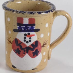 Könnte beinhalten: Eine Keramiktasse mit einem Schneemann-Design. Die Tasse hat eine beige Grundfarbe mit weißen Punkten und einem blauen Rand. Der Schneemann hat einen rot-weißen Hut, einen schwarzen Zylinder, eine rote Fliege und eine Karottennase.