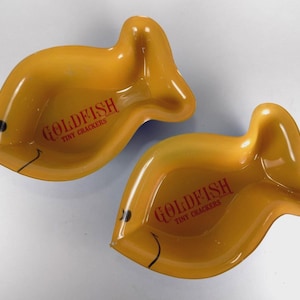 Puede incluir: Dos platos de color amarillo anaranjado con forma de pez dorado. Cada plato tiene las palabras "GOLDFISH TINY CRACKERS" impresas en rojo. Los platos tienen un ojo negro y una línea negra curva para representar una boca.