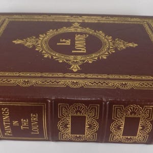 May include: A hardcover book titled "Paintings in The Louvre" with a burgundy cover and gold embossed lettering and decorative elements. The book has a gold-colored bookmark.