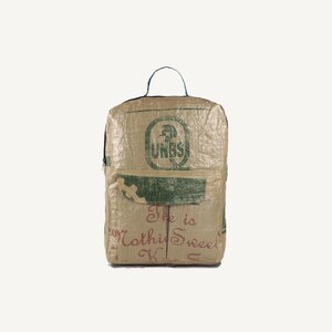 Könnte beinhalten: Beigefarbener Rucksack aus gewebtem Material mit grüner Tasche und oberem Griff. Der Rucksack hat einen Reißverschluss und ein grünes Logo. Auf der Vorderseite steht "She is Nothing Sweet".