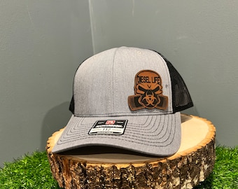 Peut inclure: Un chapeau de camionneur gris et noir avec un patch en cuir marron représentant un crâne avec un masque à gaz et le texte "Diesel Life".