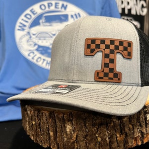 May include: A gray and black trucker hat with a brown leather patch featuring a checkered "T" on the front. The hat is sitting on a wooden surface.
