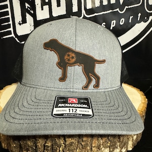 May include: A gray and black trucker hat with a brown leather patch of a dog with a Tennessee state logo. The hat has the text "Richardson" and "112 Trucker" on the front.