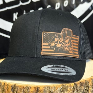 May include: Black trucker hat with a brown leather patch featuring a welder with an American flag in the background. The patch reads "The Authentic Snapback Yupoong Since 1974".