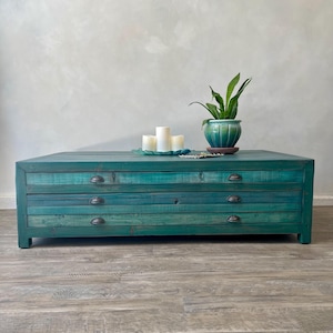 May include: A teal-colored, rectangular coffee table with three drawers and metal handles. The table has a distressed finish. On top, there are three white candles on a glass plate and a potted plant.