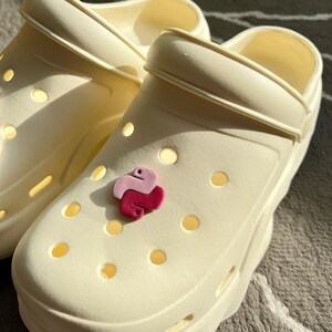 Puede incluir: Un par de zuecos blancos con un amuleto de pit&oacute;n rosa y morado sujeto a uno de los zuecos.