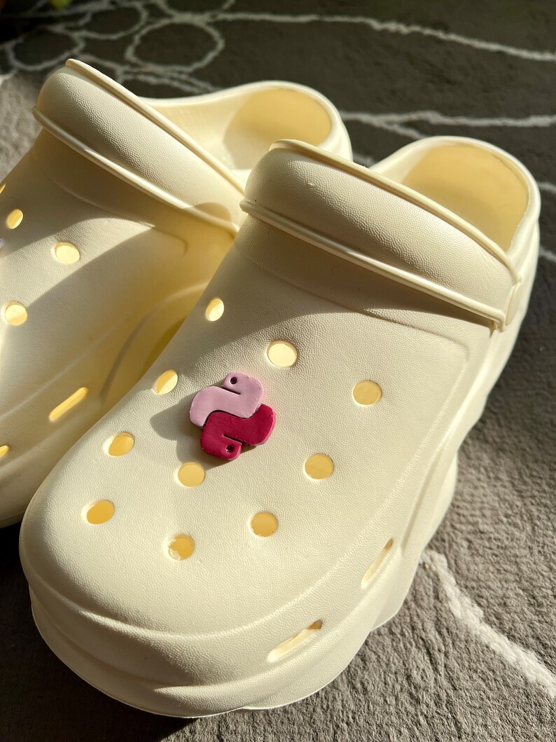 Puede incluir: Un par de zuecos blancos con un amuleto de pit&oacute;n rosa y morado sujeto a uno de los zuecos.