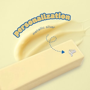 May include: A rectangular pale yellow block with a metallic silver script name tag reading Gia attached to the surface. The text personalisation appears in blue above a smear of pale yellow cream, with a dashed blue arrow pointing to the name tag.