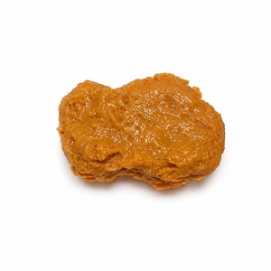 May include: A single, golden-brown chicken nugget, with a textured surface, sits against a stark white background. The nugget's irregular shape and color suggest it is freshly cooked and ready to eat. The image is a studio shot.