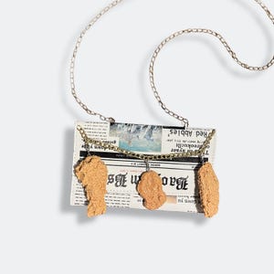 Può includere: Una borsa a mano originale con un design a stampa di giornale, con tre ciondoli a forma di pollo fritto. La borsa ha una tracolla a catena argentata e un dettaglio a catena dorata. La stampa del giornale include il testo "Bao B" e "Red Abbies".