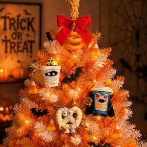 May include: An orange and white artificial tree decorated for Halloween with a fried chicken topper, a red bow, candy corn ornaments, black bats, a mummy figure, a pretzel with eyes, and a blue coffee cup ornament with a vampire graphic.