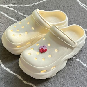 Puede incluir: Un par de zuecos blancos de plataforma con una superficie texturizada y un peque&ntilde;o amuleto morado y rojo sujeto a uno de los zuecos.