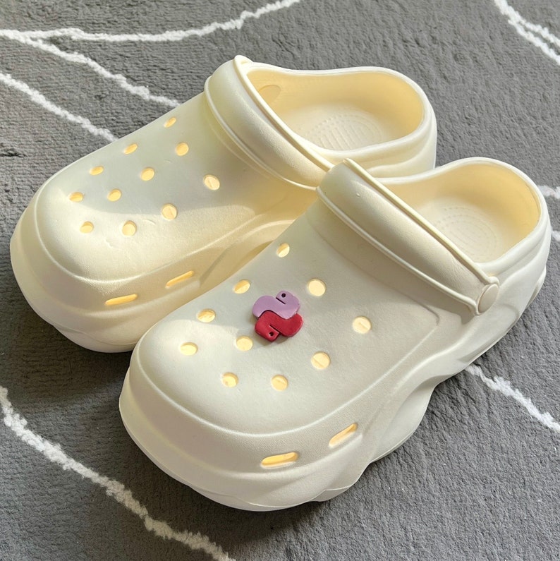 Puede incluir: Un par de zuecos blancos de plataforma con una superficie texturizada y un peque&ntilde;o amuleto morado y rojo sujeto a uno de los zuecos.