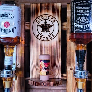 Puede incluir: Un dispensador de licores de madera con dos botellas de whisky, Jim Beam y Jack Daniel's, y un vaso de chupito. El dispensador tiene un logotipo de los Astros de Houston y una peque&ntilde;a repisa con un vaso de chupito decorativo. La madera tiene un acabado r&uacute;stico y quemado.