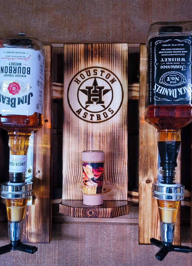 Puede incluir: Un dispensador de licores de madera con dos botellas de whisky, Jim Beam y Jack Daniel's, y un vaso de chupito. El dispensador tiene un logotipo de los Astros de Houston y una peque&ntilde;a repisa con un vaso de chupito decorativo. La madera tiene un acabado r&uacute;stico y quemado.