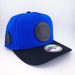 Peut inclure: Casquette de baseball bleue avec une visière noire. Le chapeau présente un emblème circulaire avec le texte "DEPORTIVO CRUZ AZUL MEXICO" et un symbole de croix. Un petit emblème est sur le côté.
