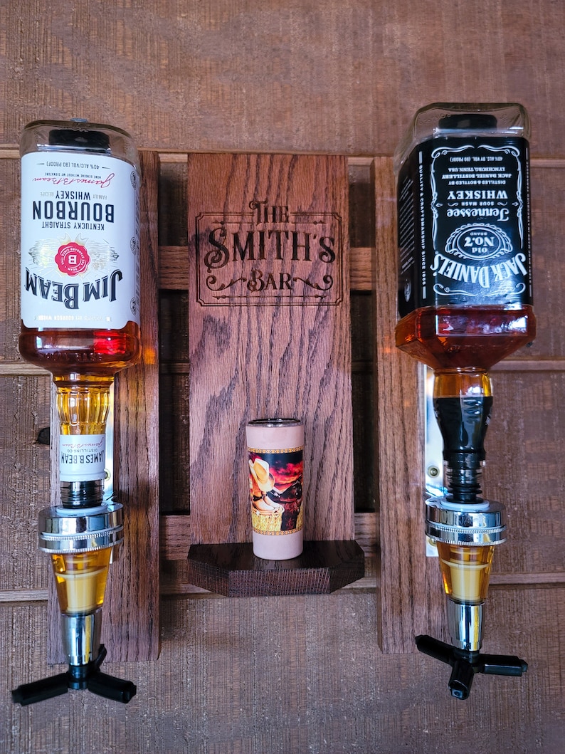 Puede incluir: Un expositor de bar de madera con dos dispensadores de botellas de licor, uno para Jim Beam Bourbon y otro para Jack Daniel's Tennessee Whiskey. Una peque&ntilde;a repisa contiene un objeto decorativo, y el letrero de madera dice "The Smith's Bar."