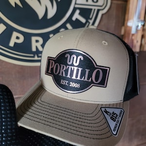 Puede incluir: Una gorra de béisbol de color beige y negro con un parche bordado en negro y oro rosa que dice "JC Portllo Est. 2008".