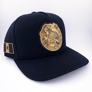 Op de afbeelding: Een marineblauwe baseballcap met een gouden embleem van het Mexicaanse wapen aan de voorkant. De cap heeft ook een klein gouden embleem van de Mexicaanse vlag aan de zijkant. De cap heeft een gebogen klep.