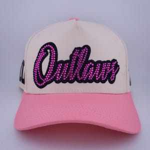 Op de afbeelding: Een baseballpet met een crèmekleurige kroon en een effen roze klep. Op het voorpaneel staat het woord Outlaws in een cursief lettertype, omlijnd in het zwart en gevuld met kleine, sprankelende roze strass-steentjes die het licht vangen.