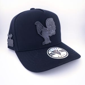 Op de afbeelding: Zwarte baseballcap met een grijs hanenembleem. De cap heeft een gebogen klep en een gestructureerde kroon. De merknaam "DE LEON" is zichtbaar op een sticker op de klep. De cap heeft ook de tekst "ANGELO FINO" aan de zijkant.