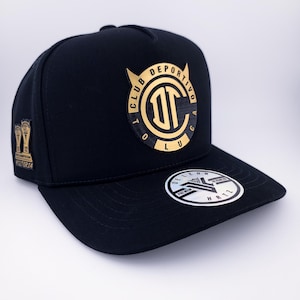 May include: Black baseball cap featuring a gold emblem with the text "CLUB DEPORTIVO TOLUCA" and a stylized "T" logo. The cap has a flat brim and a small pin on the side. A sticker is on the brim.