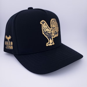 Op de afbeelding: Zwarte baseballcap met een gouden haan op de voorkant en een kleiner haanembleem aan de zijkant. De tekst "GALLO FINO" staat onder het zijembleem. De cap heeft een gebogen klep en een knoop aan de bovenkant.