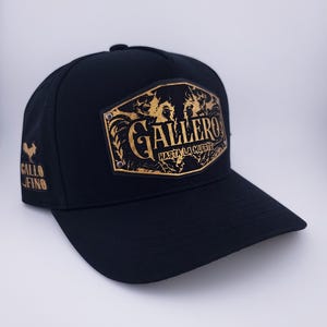 Op de afbeelding: Zwarte baseballcap met een gebogen klep. De cap heeft een gouden embleem met het woord "GALLERO" en de zin "HASTA LA MUERTE". Een gouden haanlogo en de woorden "GALLO FINO" staan op de zijkant.