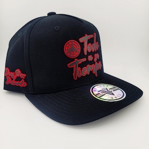 Op de afbeelding: Zwarte baseballcap met rode geborduurde tekst en logo. De cap heeft de woorden "Todo a su Tiempo" en "Dios Por Delante". Een holografische sticker zit op de rand.