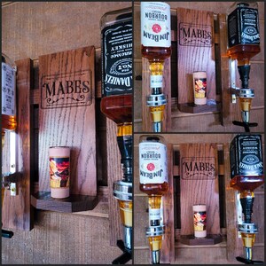 Puede incluir: Un dispensador de licores de madera con el nombre "MABES" grabado. El dispensador contiene botellas de whisky bourbon Jack Daniel's y Jim Beam. Un peque&ntilde;o vaso de chupito est&aacute; en un estante debajo.