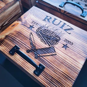May include: A wooden box with a black metal handle. The box is engraved with the text "RUIZ EST. 2003" and a star on each side of the text. The box also has an image of a steak on fire with a knife.