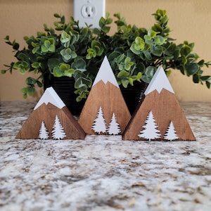May include: Three wooden triangle-shaped mountains with white snow peaks and white silhouette trees. The mountains are arranged in a row from largest to smallest.