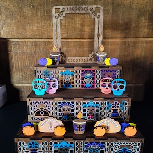 May include: A wooden Day of the Dead altar with multiple levels, adorned with colorful skull figurines, candles, and decorative cutouts. The altar features a central frame and is decorated with vibrant orange, blue, pink, and purple accents. The overall theme is a celebration of the Day of the Dead.