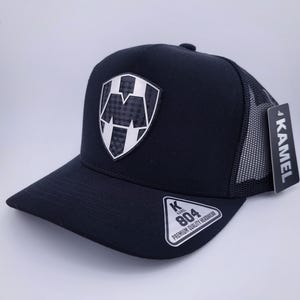 Peut inclure: Casquette de baseball noire et blanche avec un dos en mesh. La casquette arbore un logo argenté et noir avec la lettre "M" à l'intérieur d'un bouclier. La casquette comporte également une étiquette noire et blanche avec le texte "K Kamel 804 Premium Quality Headwear".