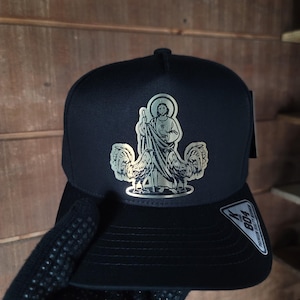 Peut inclure: Casquette de baseball noire avec un patch métallique argenté représentant une figure religieuse et deux dindes. Le patch a une bordure argentée et le texte "804" est visible sur le côté de la casquette.