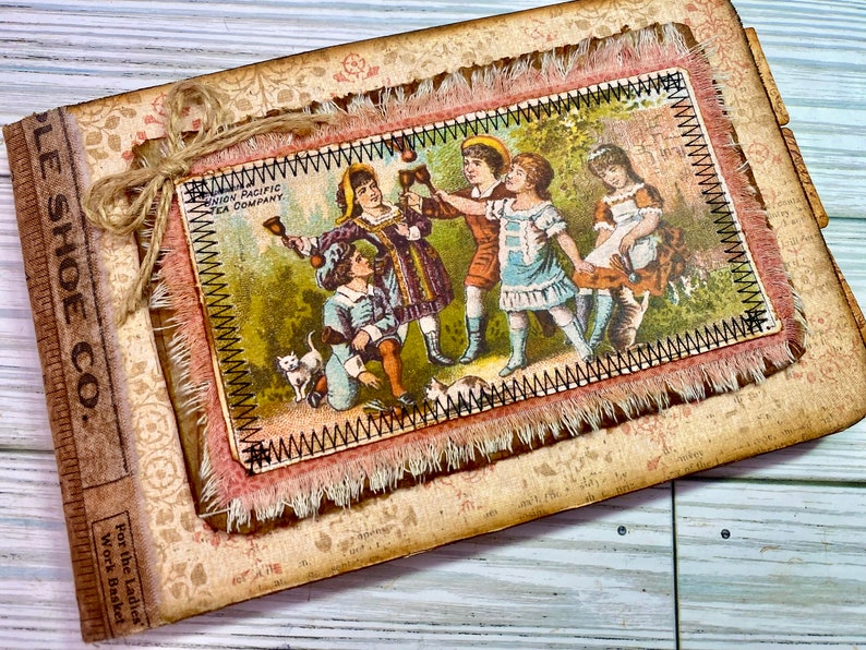 Pu&ograve; includere: Un diario in stile vintage con una copertina in pelle marrone e una patta in tessuto decorativa. La patta presenta un bordo cucito e un bordo sfrangiato. Il diario ha un titolo sulla copertina che recita "The Shoe Co.". La patta presenta un'illustrazione vintage di bambini che giocano con i campanelli. L'illustrazione &egrave; della "Union Pacific Tea Company".