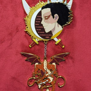May include: Gold and red enamel pin featuring a character with black hair and a red mark on their face. The character is surrounded by a sun and moon design. The pin also features a dragon with red and gold wings. The text on the pin reads: "No one can give you your honour. It is something you earn for yourself. By choosing to do what is right."