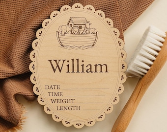 May include: An oval wooden plaque with a scalloped edge featuring an engraved Noah's Ark illustration with animals, the name William, and blank lines for date, time, weight, and length. A wooden baby hairbrush is positioned next to the plaque.